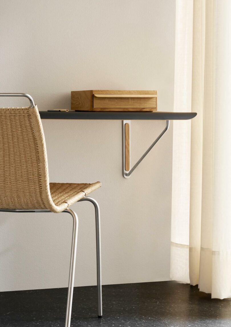 A Carl Hansen & Son woven chair sits in front of a minimalist floating black shelf with a wooden box on top, next to a cream curtain