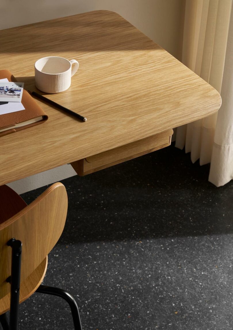 A wooden desk with a cup, notebook, pencil, and a photograph on top, next to a Carl Hansen & Son Anker Bak wooden chair and cream-colored curtains