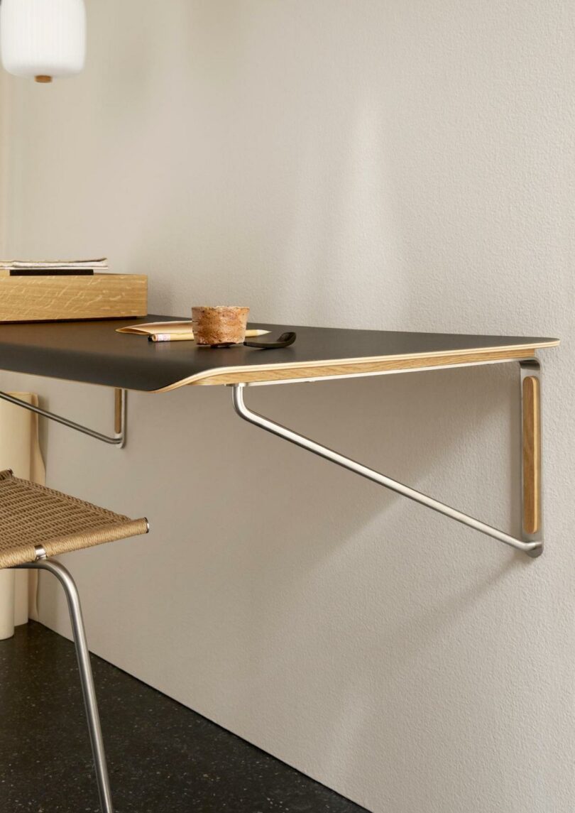 A minimalist wall-mounted desk with a black surface and metal supports, paired with a Carl Hansen & Son Anker Bak woven chair. On top sit a notebook, pen, cup, and small dish—perfect for streamlined workspaces