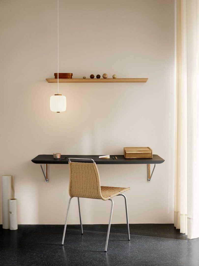 Minimalist workspace featuring a Carl Hansen & Son Anker Bak woven chair at a wall-mounted desk, a wooden shelf with decor items above, and a nearby pendant light. Natural light enters from the right