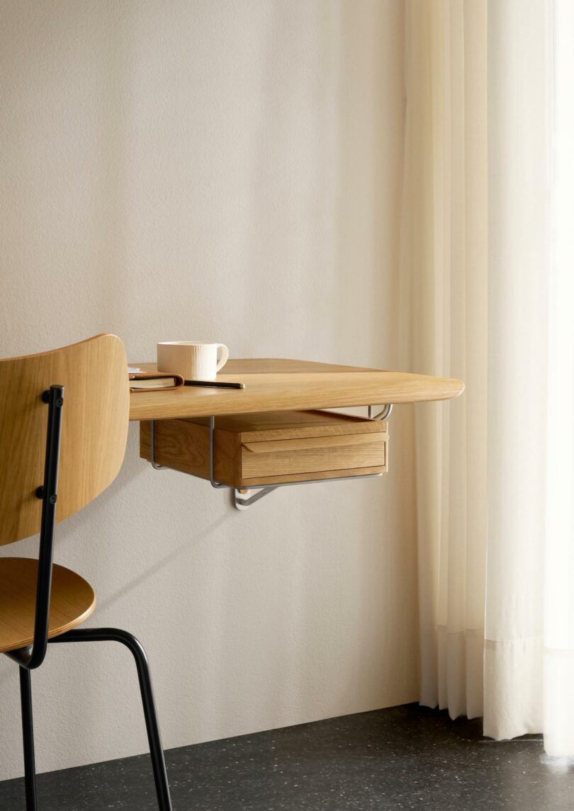 A minimalist Carl Hansen & Son wooden desk with a drawer, a matching chair by Anker Bak, a closed notebook, pen, and white mug sit by a window with sheer curtains