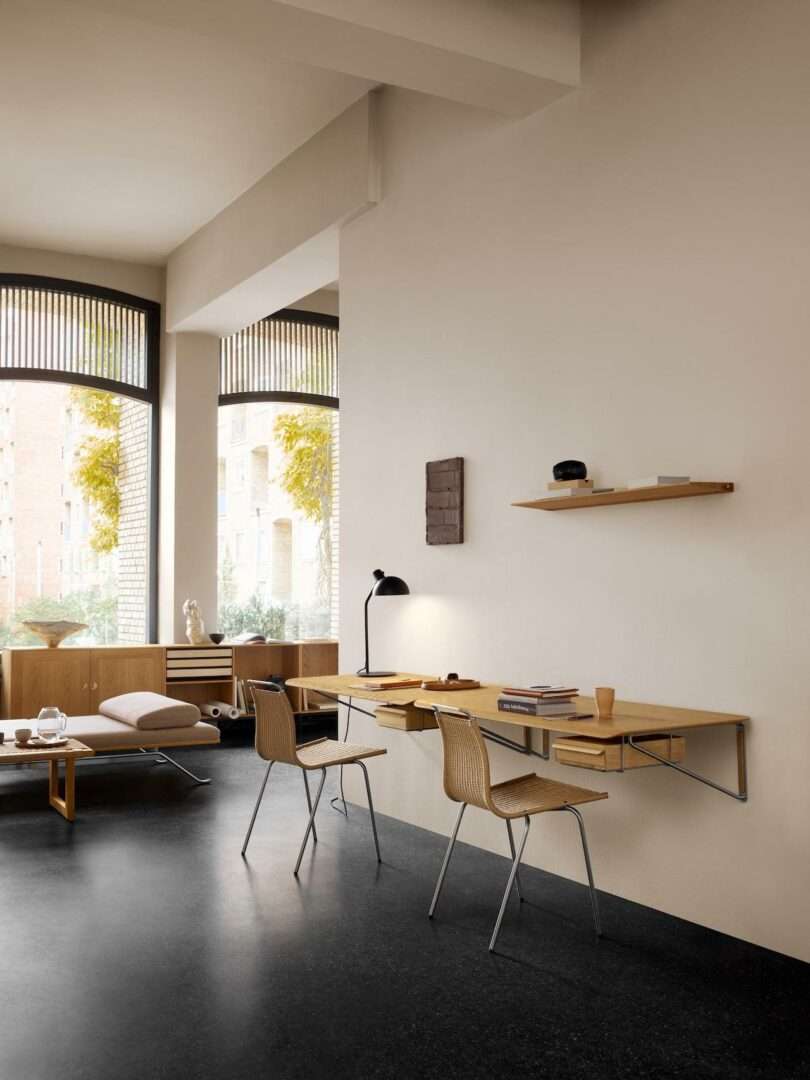 Minimalist home office with a Carl Hansen & Son Anker Bak floating wooden desk, two chairs, wall shelf, lamp, and large windows letting in natural light. Neutral color palette and simple decor