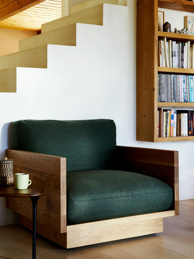 A green cushioned armchair with wooden frame sits next to a small round table and a built-in bookshelf under a modern staircase