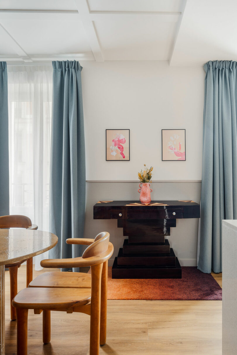 A modern dining area with wooden chairs, a round table, blue curtains, a black console table, pink artwork, and a vase with dried flowers on a red rug.