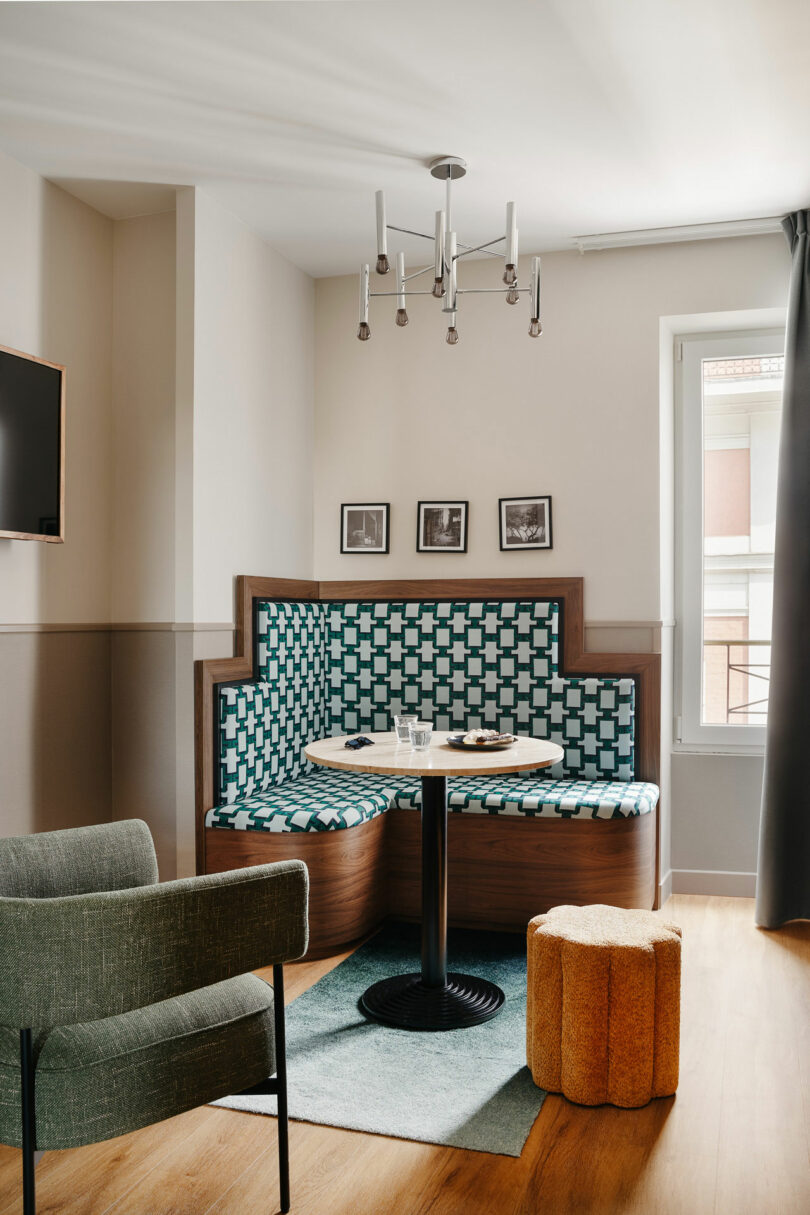 A modern seating nook with geometric-patterned upholstery, a round table, armchair, yellow pouf, wall art, and a chandelier in a bright room with wood flooring.
