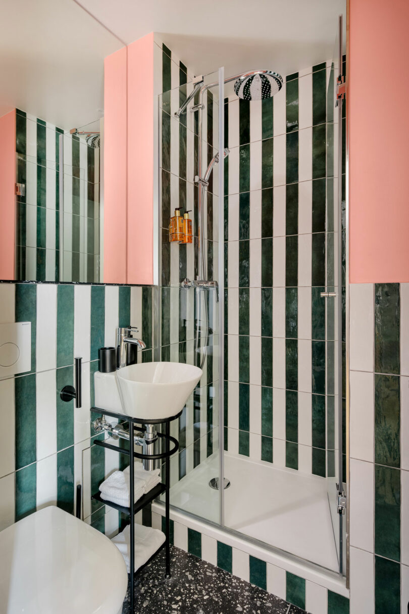 A modern bathroom with green and white vertical striped tiles, a glass shower enclosure, a white sink with black fixtures, and pink accents on the walls.