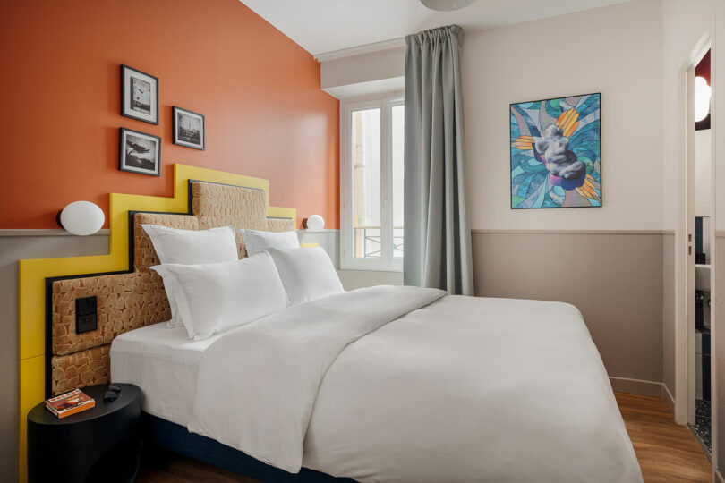 A modern bedroom with an orange accent wall, a bed with white linens, two small bedside tables, wall art, and a window with gray curtains.