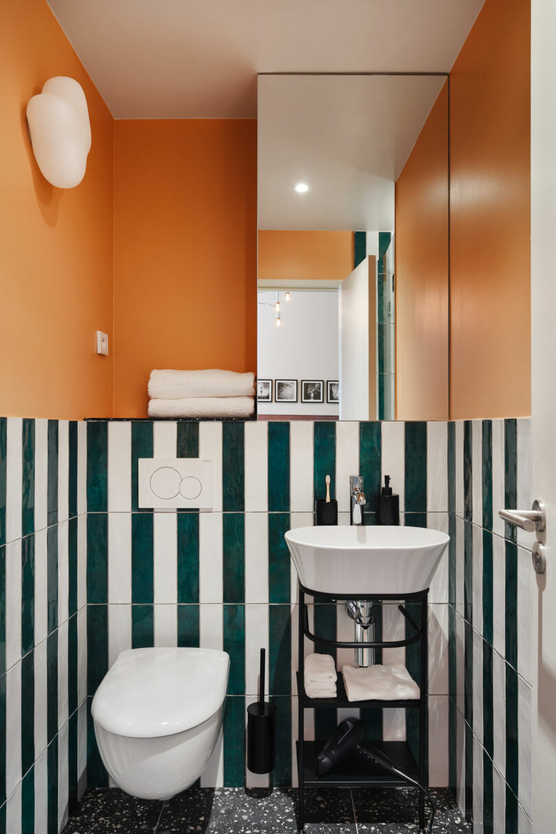 Modern bathroom with orange walls, green and white vertical striped tiles, wall-mounted toilet, small sink, mirror cabinet, and stacked towels.