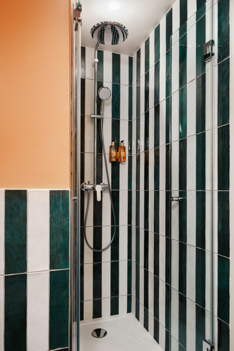 A shower stall with vertical green and white striped tiles, a rainfall showerhead, a handheld shower, and wall-mounted soap dispensers.