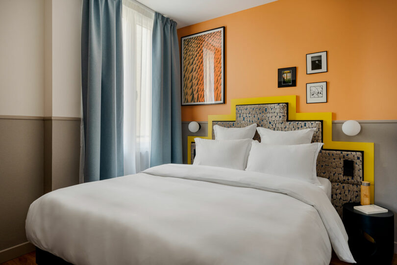 A modern hotel room with a double bed, white linens, a woven headboard with a yellow frame, orange accent wall, window with blue curtains, and framed artwork.