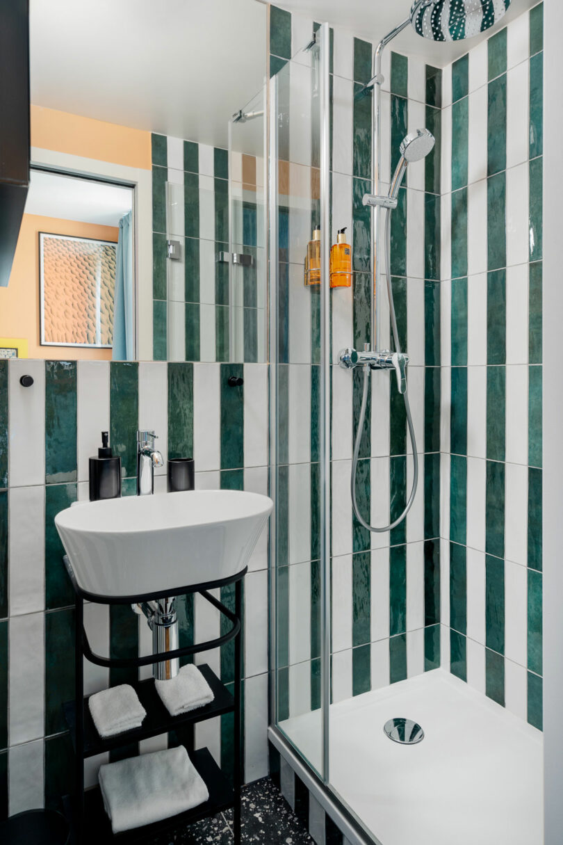 Modern bathroom with green and white vertical striped tiles, a glass shower enclosure, wall-mounted showerhead, sink with black fixtures, and shelves holding towels and toiletries.