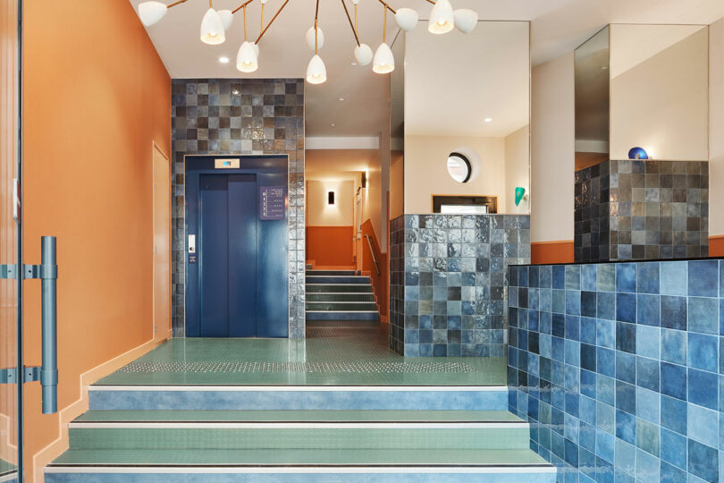 Modern building lobby with green tiled steps, blue tiled walls, a blue elevator, and contemporary ceiling light fixture.
