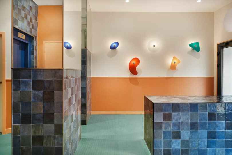 Brightly lit interior with colorful wall lamps, blue and brown tiled surfaces, green flooring, and an elevator door on the left.