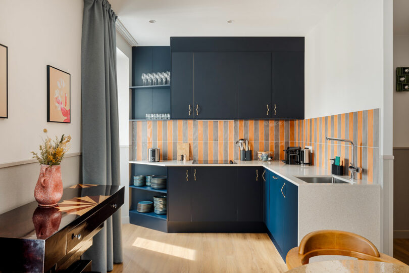 Modern kitchen with dark blue cabinets, orange-striped backsplash, open shelves with glassware, a dining counter, and light wood flooring.