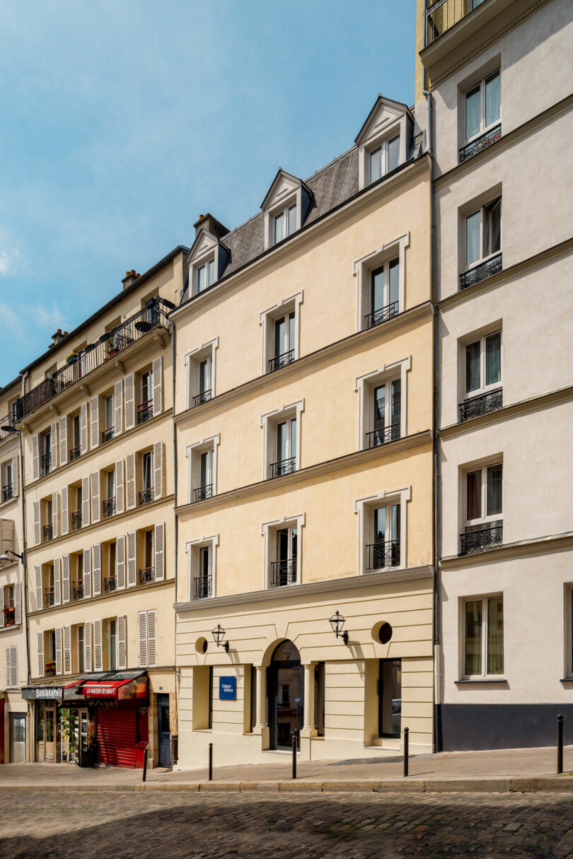 A beige and yellow mid-rise apartment building with dormer windows on a cobblestone street, adjacent to a red storefront.