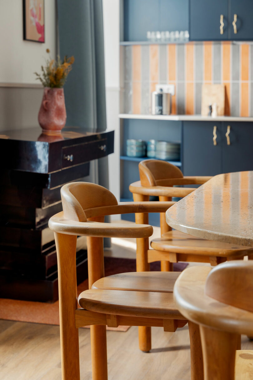 A close-up view of wooden dining chairs and a table in a modern kitchen with blue cabinets and patterned backsplash in the background.