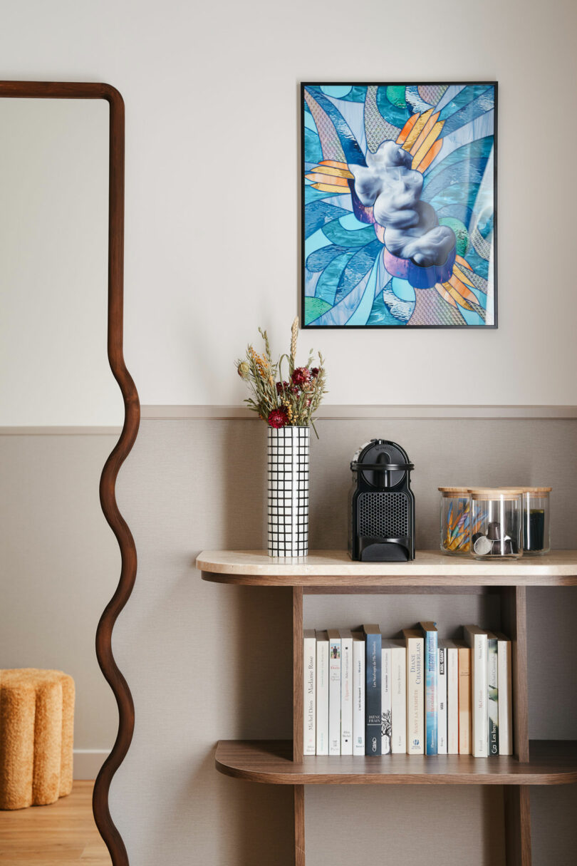 A modern console table with books, a vase of dried flowers, a coffee machine, and a glass jar, beneath a colorful abstract framed artwork.