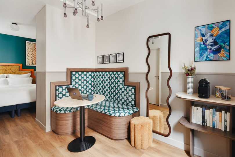 A modern hotel room seating area with a geometric-patterned bench, small round table, wall mirror, bookshelf, coffee maker, and a glimpse of a bed in the adjoining room.