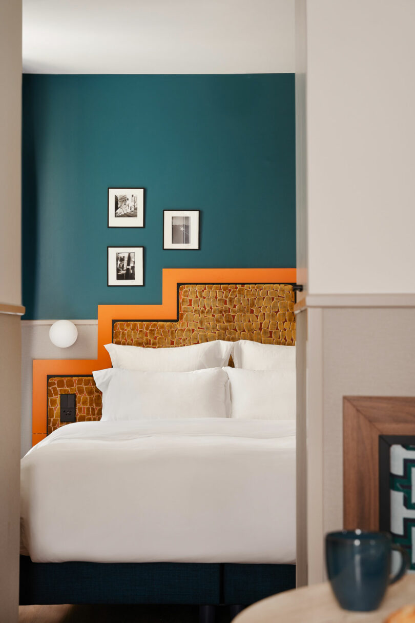 A neatly made bed with white linens, a textured orange headboard, and three framed photos on a teal wall in a modern hotel room.