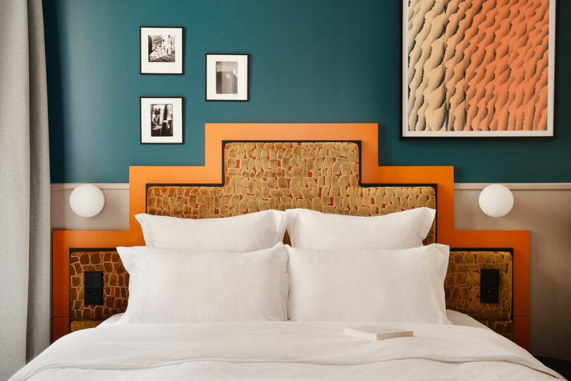 A neatly made bed with white linens, an orange and textured headboard, framed artwork on a teal wall, and a closed book on the duvet.