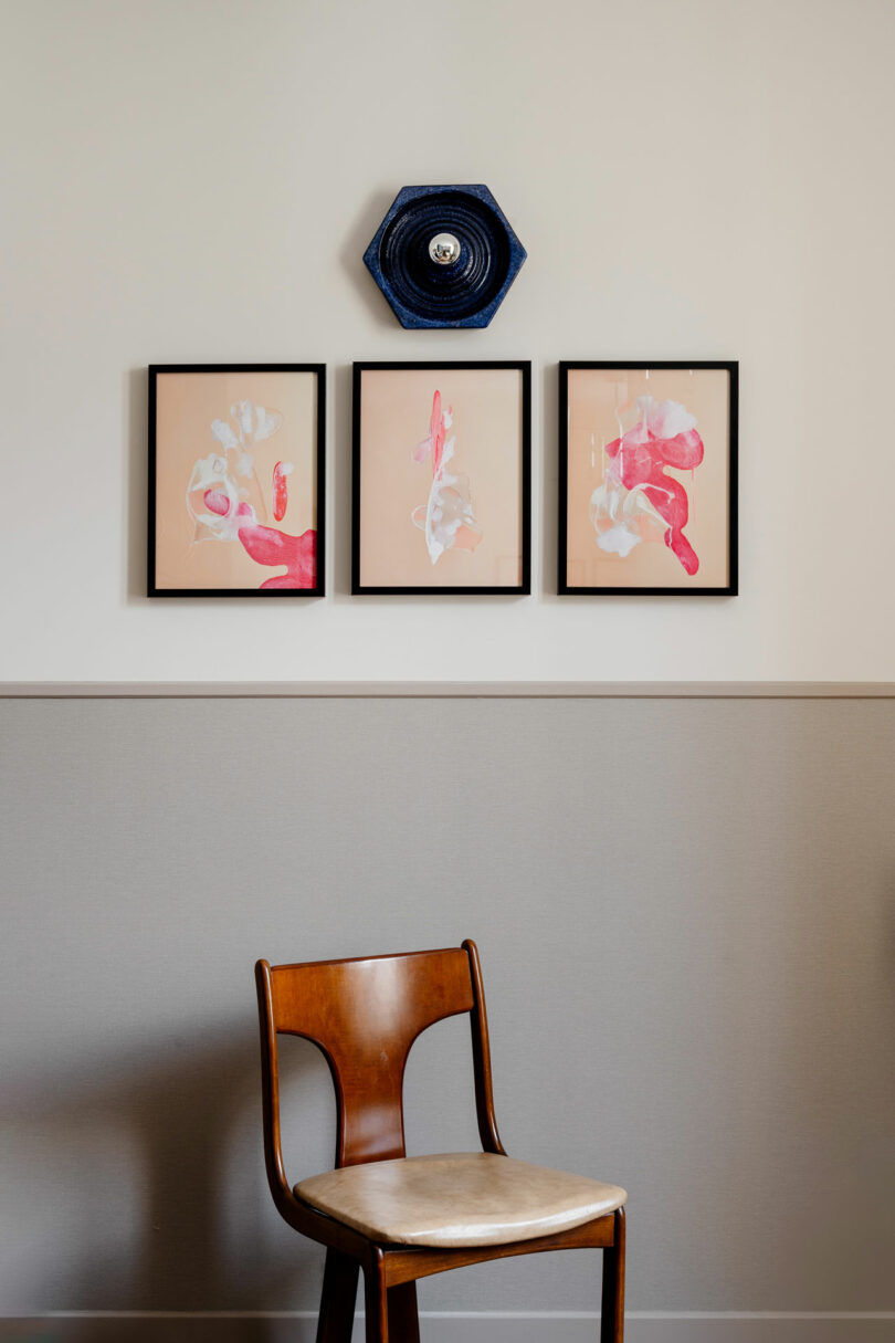 A wooden chair sits in front of a beige wall, above which are three framed abstract artworks and a blue ceramic wall decoration.
