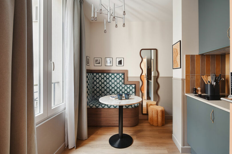 Modern dining nook with patterned banquette, marble-top table, large mirror, decorative light fixture, and kitchen area with blue cabinets and brown tile backsplash.