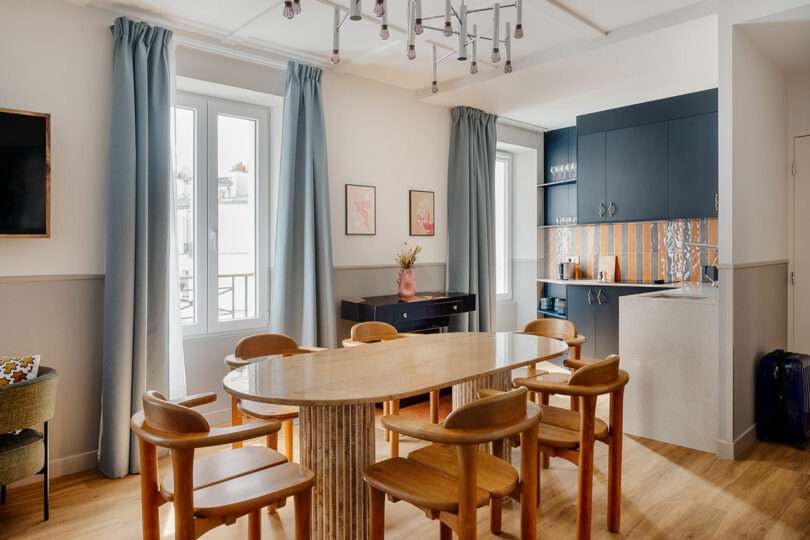 A modern dining area with a light wood table, eight wooden chairs, blue curtains, and an open kitchen with dark cabinets and a patterned backsplash.