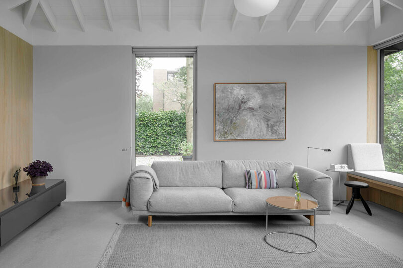Minimalist living room with a light gray sofa, round coffee table, abstract painting on the wall, large windows, and neutral decor.