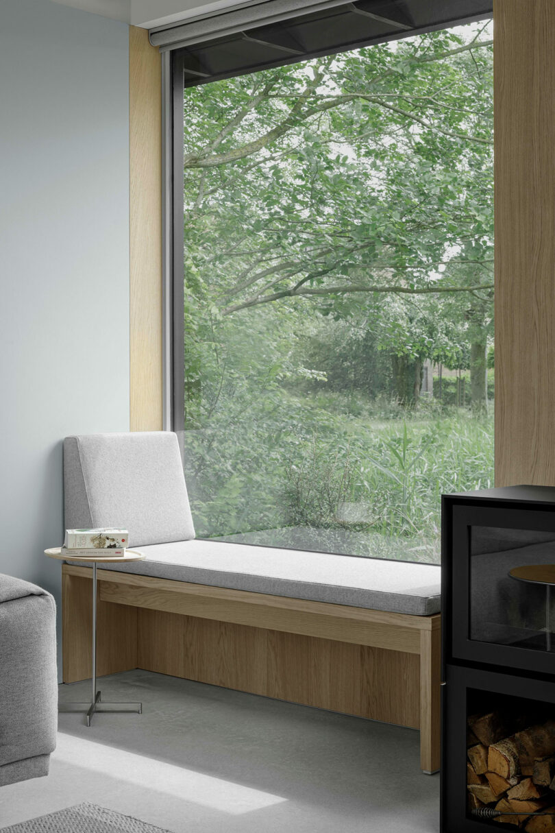 A modern window seat with a light gray cushion is set beside a large window overlooking green trees, next to a black wood stove with firewood stored below.