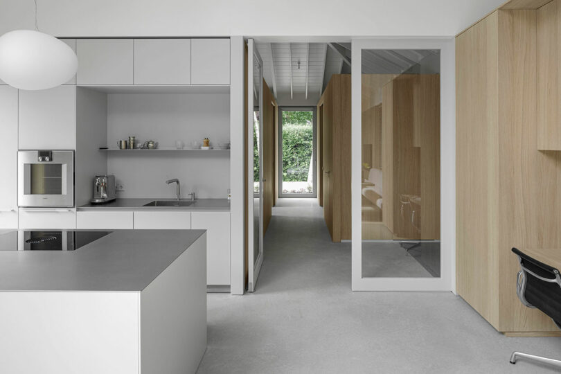 Minimalist kitchen with gray cabinets and island, stainless steel appliances, and a hallway with glass and wood paneling leading to a green outdoor area.