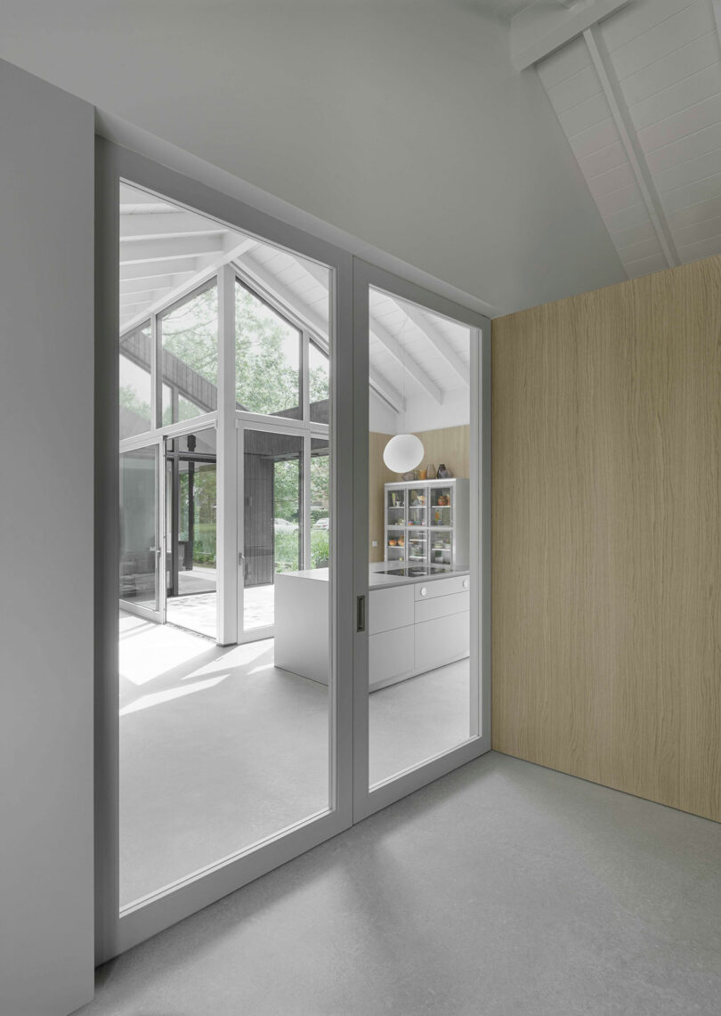 A modern interior with sliding glass doors opens to a bright kitchen area and a view of trees outside; neutral colors and clean lines dominate the design.