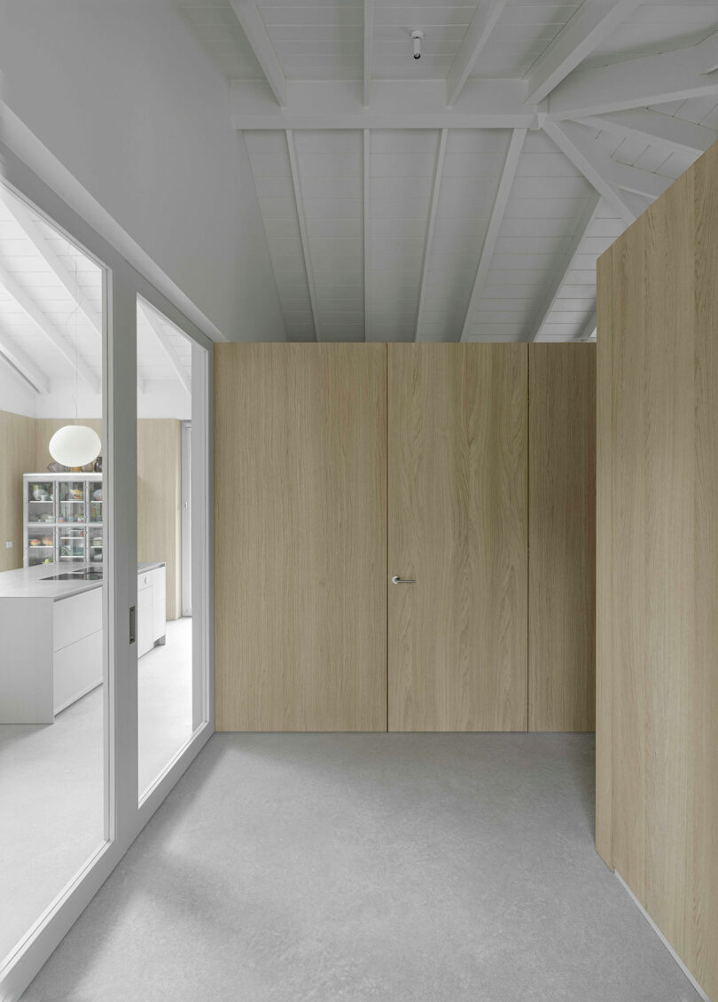 Minimalist interior with light wood panel walls, grey floor, white ceiling beams, and large glass window showing a modern kitchen area with shelves and a round ceiling light.