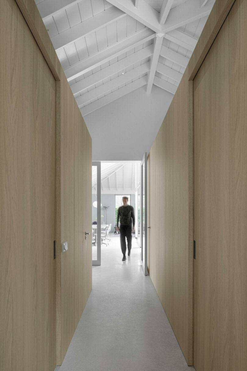 A person walks away down a hallway with light wood walls and a vaulted white ceiling, toward a bright room with large windows.