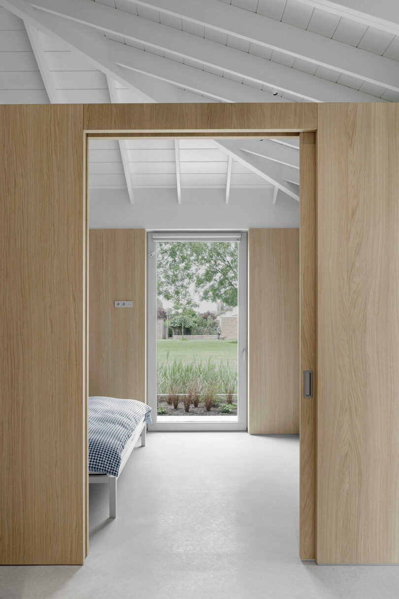Minimalist bedroom with light wood sliding doors, white ceiling beams, and a bed with a blue checkered blanket. A glass door at the far end opens to a garden with greenery outside.