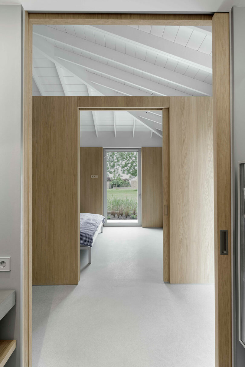 View through open wooden sliding doors into a modern minimalist bedroom with white ceiling beams, a bed, large window, and garden visible outside.