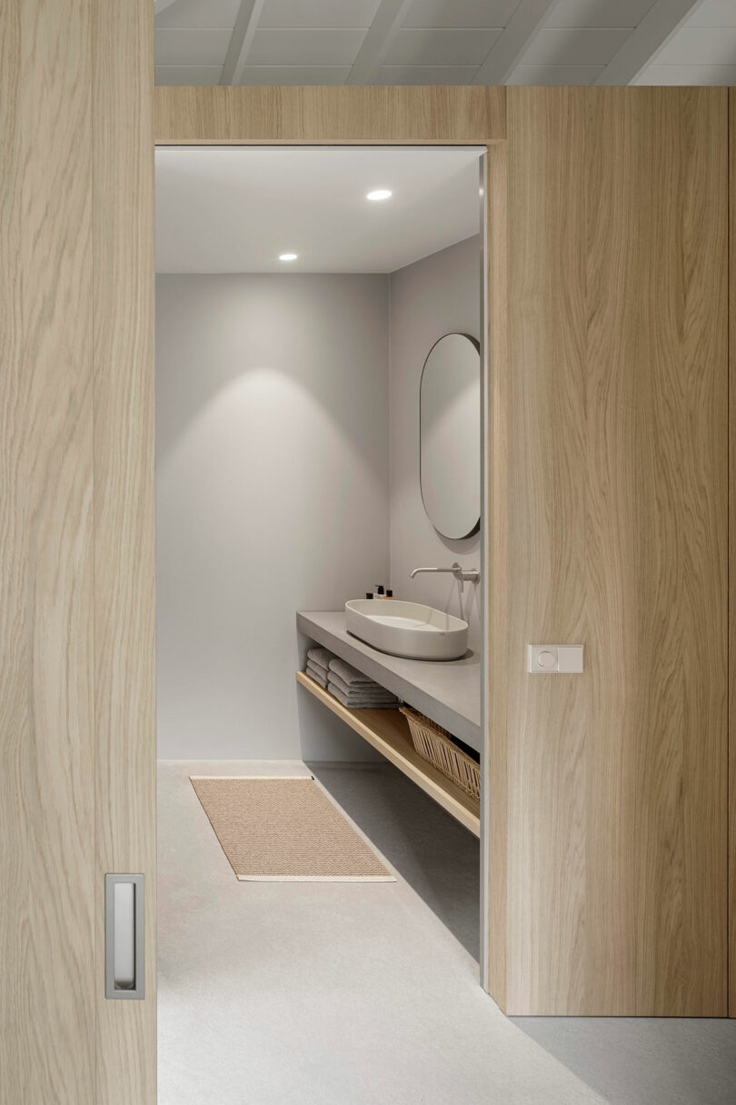 A modern bathroom with light wood sliding door, oval mirror, vessel sink, open shelf with towels and baskets, gray walls, and recessed ceiling lights.