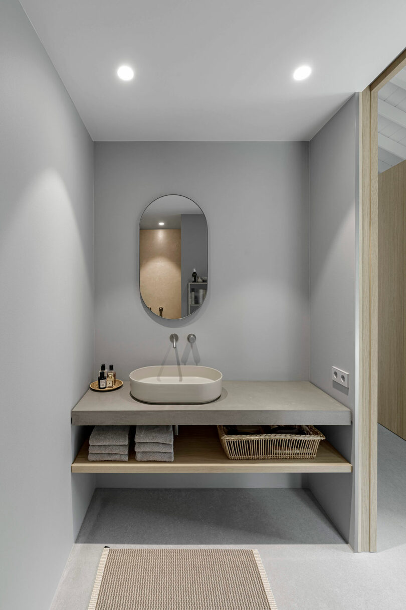 Minimalist bathroom with a round mirror, oval sink, wall-mounted faucets, toiletries, folded towels, a wicker basket, and neutral grey walls under recessed lighting.