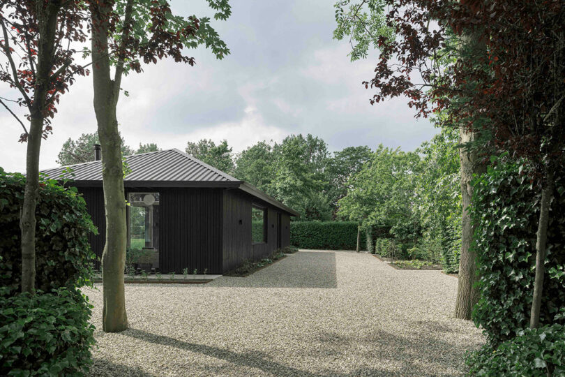A modern black wooden house with a sloped metal roof sits in a gravel yard, surrounded by green trees and hedges under a cloudy sky.
