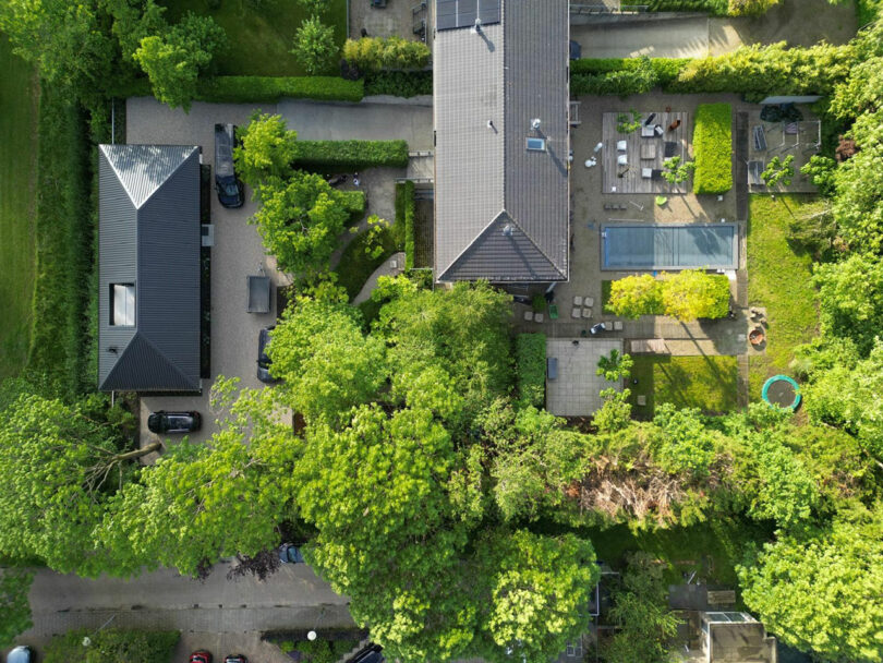 Aerial view of a residential property with two buildings, a swimming pool, patio areas, parked cars, and surrounding greenery.