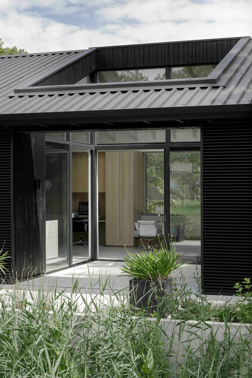 Modern black house with large glass doors and windows, open to a minimalist interior; tall grass and potted plant in the foreground.