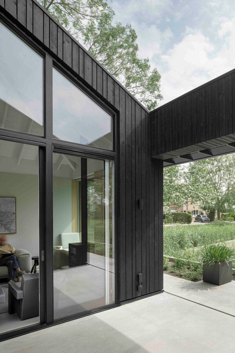 Modern house exterior with large glass windows and black wood cladding, showing an indoor seating area and outdoor greenery under a partly cloudy sky.