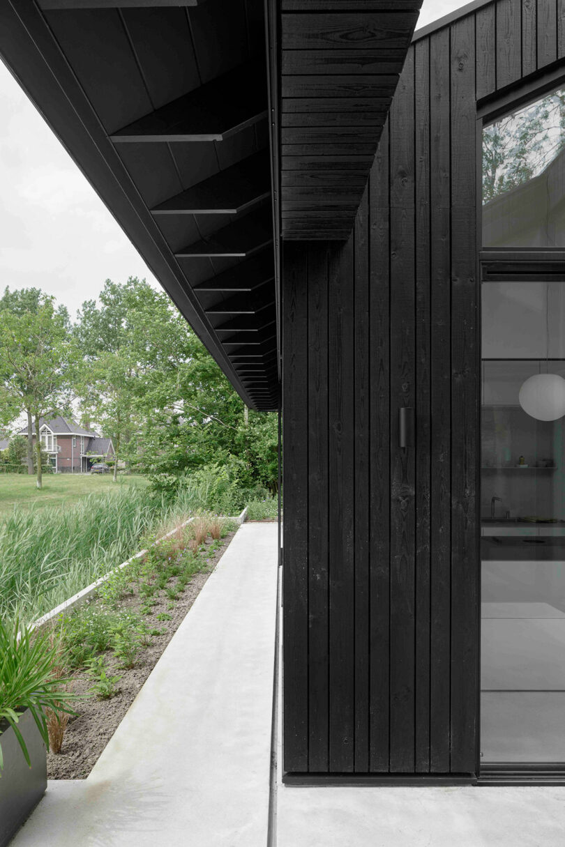 A modern black wooden house exterior with vertical paneling, large windows, a concrete walkway, and green landscaping on a cloudy day.