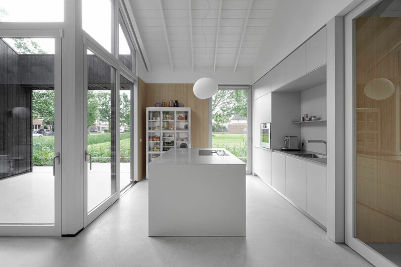 Modern kitchen with large windows, a central island, open shelving, minimalist white cabinets, and views of greenery outside.