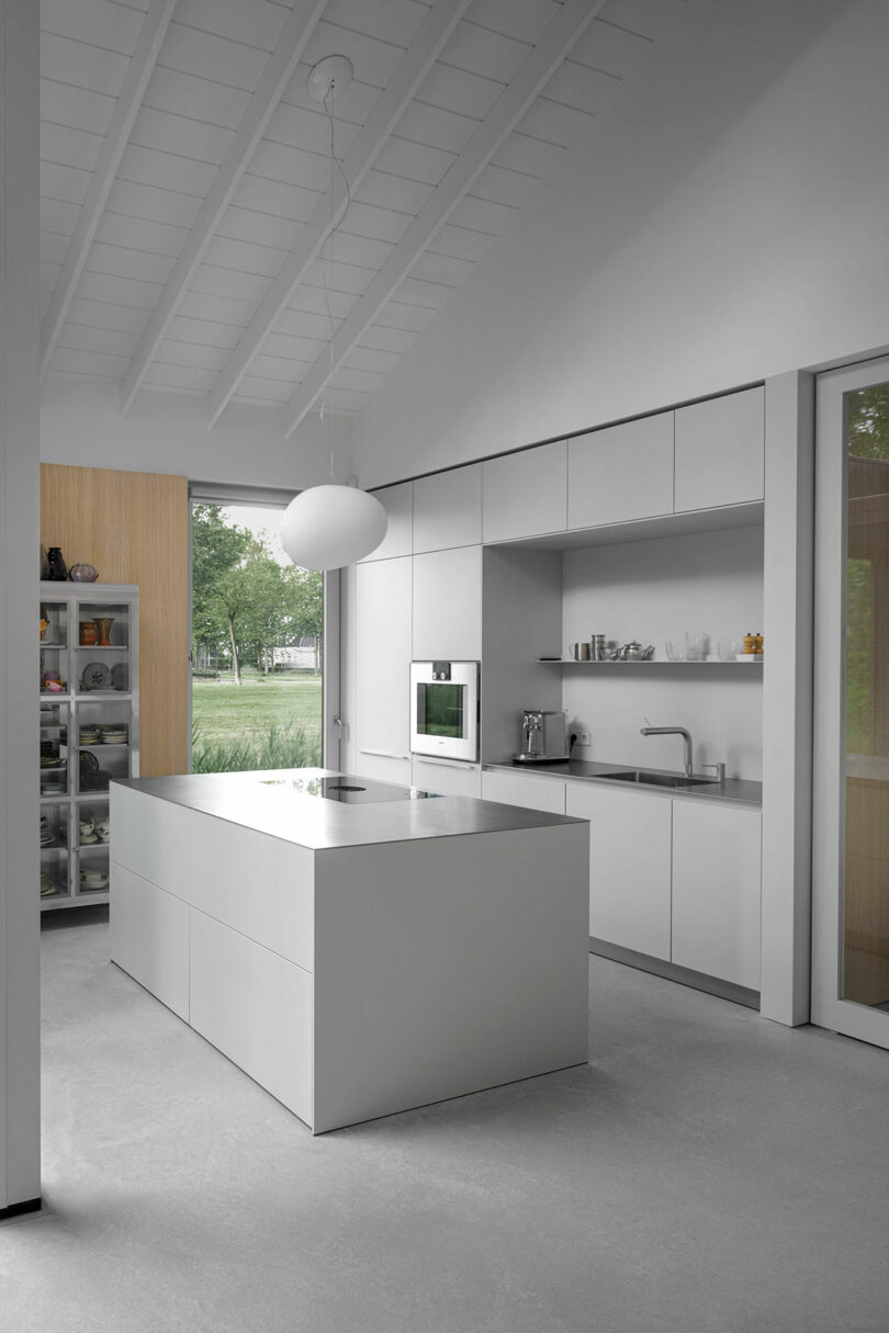 Minimalist modern kitchen with white cabinetry, an island, built-in appliances, open shelving, large window, and high sloped ceiling with exposed beams.