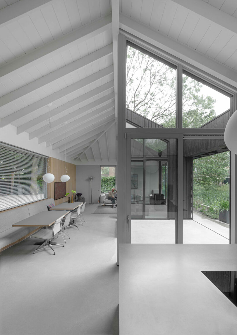 Modern open-plan interior with white walls and ceilings, large glass windows, a dining area with white chairs, and a view of greenery outside.