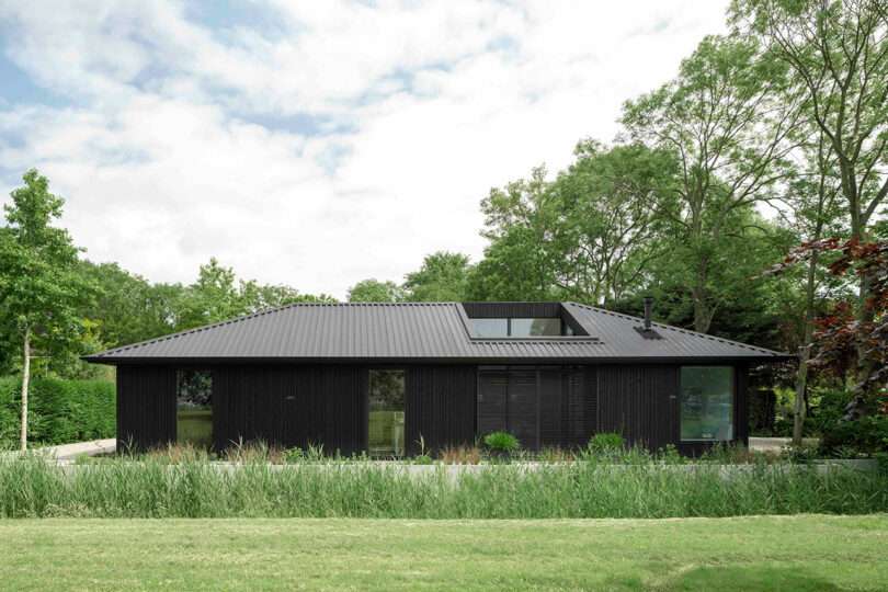 A modern, single-story house with a black exterior and sloped roof, surrounded by trees and greenery under a partly cloudy sky.