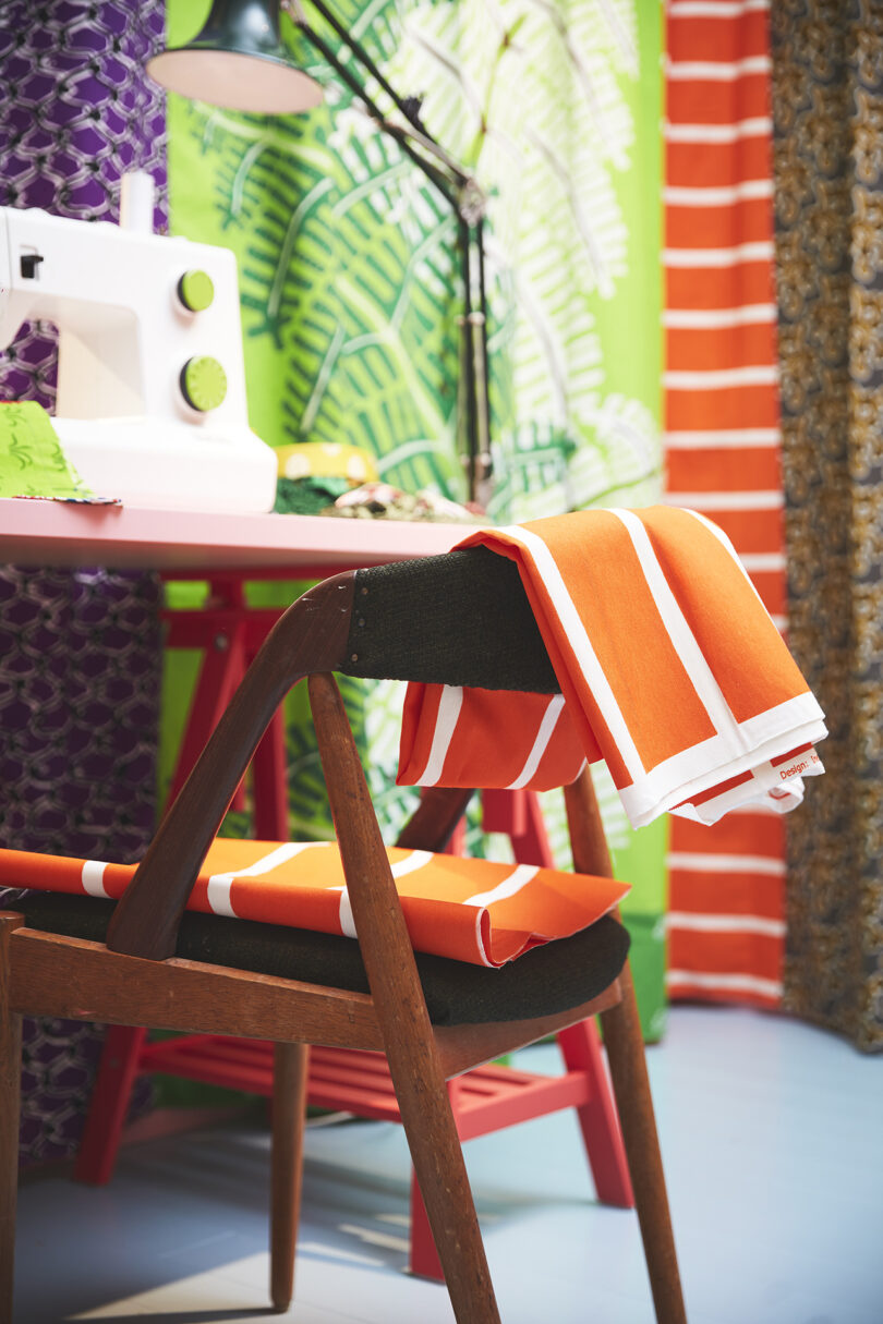 A sewing machine on a pink desk with a wooden chair draped in orange and white striped fabric, surrounded by colorful patterned curtains.