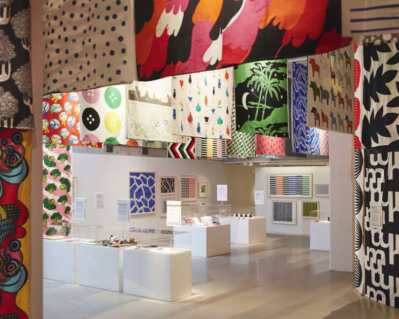 A bright exhibition space displays various colorful textiles hanging from the ceiling, with art pieces and informational panels arranged on white pedestals and walls.