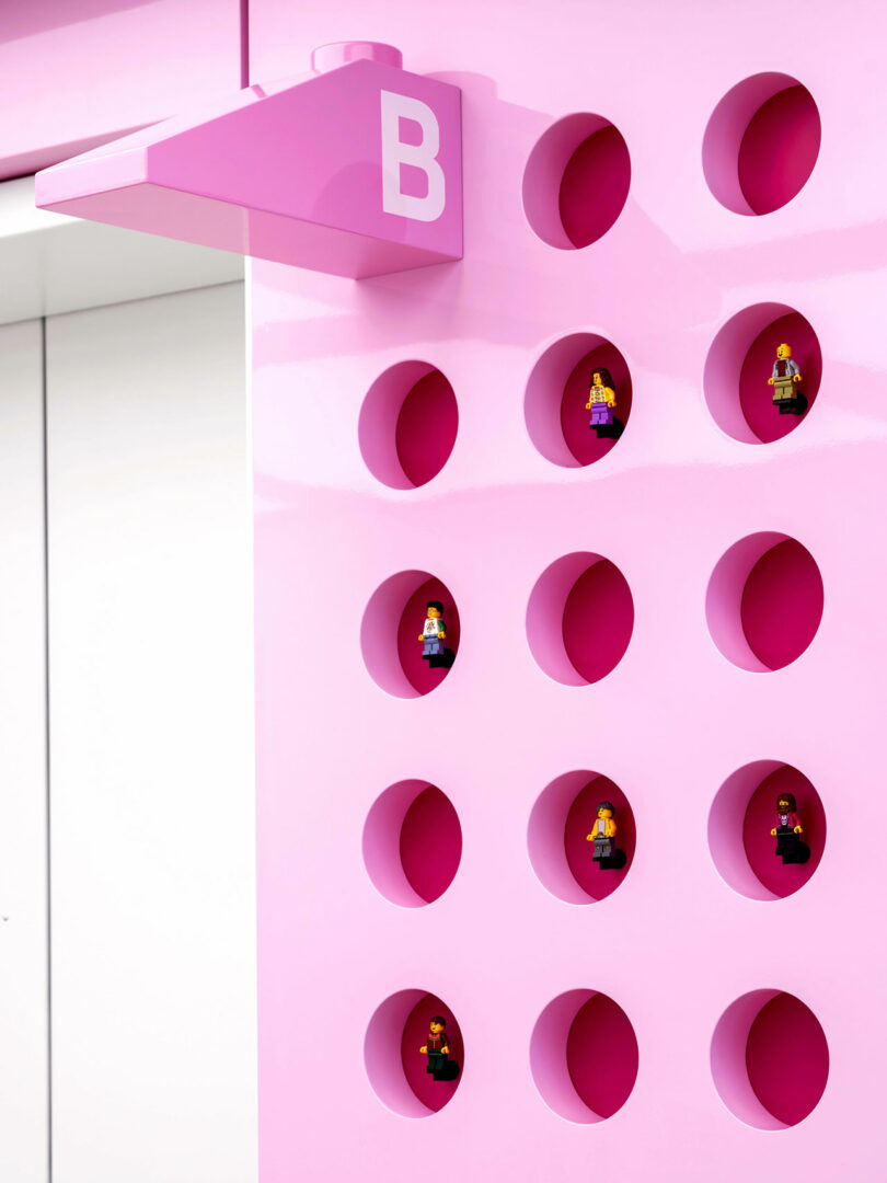 A pink wall with circular holes, each showcasing a LEGO minifigure, sits beside a pink sign labeled 