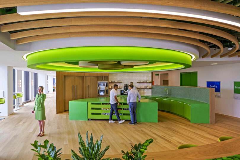 Modern office kitchen with a bright green circular ceiling fixture, three people conversing near LEGO-green counters, and one person standing separately; plants and seating visible.